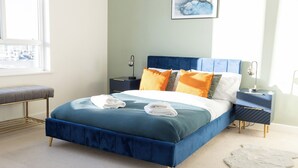A modern bedroom with a blue bed, a gray bench, and a nightstand with a lamp. - Stylish 2BR Apartment, Town Centre, Free Parking (Hatfield)