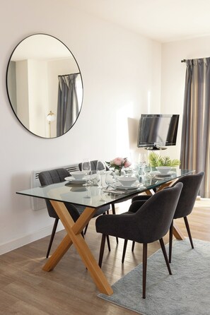 Un espace repas moderne avec une table en verre, des chaises foncées, un miroir rond et un téléviseur à écran plat.
