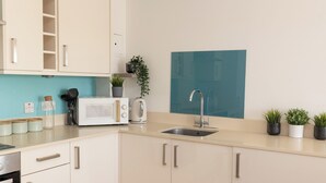 A modern kitchen with white cabinets, a stainless steel sink, a microwave, and a toaster. There are potted plants on the counter and a blue backsplash. - Stylish 2BR Apartment, Town Centre, Free Parking (Hatfield)