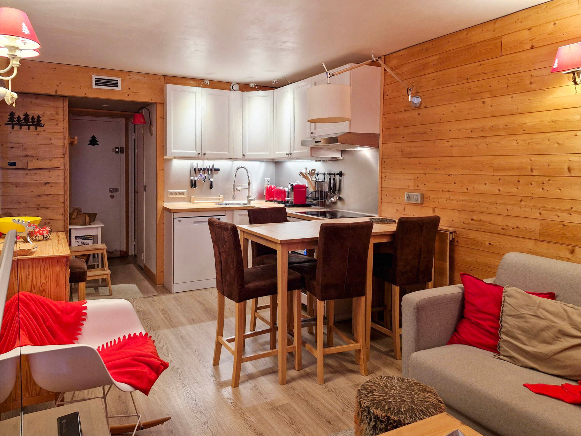 A compact kitchen with white cabinets, a dining table with four chairs, and a sofa with red cushions.