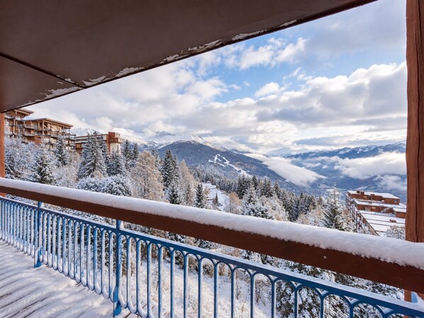 Un balcón con barandilla cubierta de nieve, con vistas a un paisaje montañoso con edificios y árboles.