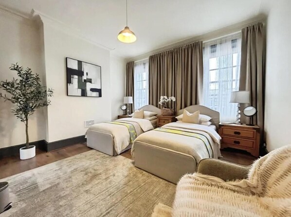 A hotel room with two beds, a large window with curtains, a potted plant, a painting, and a lamp on a bedside table.