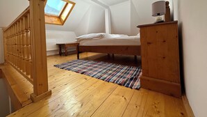 A bedroom with a wooden bed, a small bedside table, a nightstand, a window with a view, a ceiling light, and a wooden staircase. - Cazare Apuseni  Pensiune cu ciubăr (Vidra)
