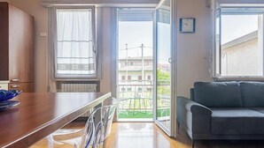 A room with a wooden table, chairs, a sofa, and a sliding glass door leading to a balcony with a view of buildings.
