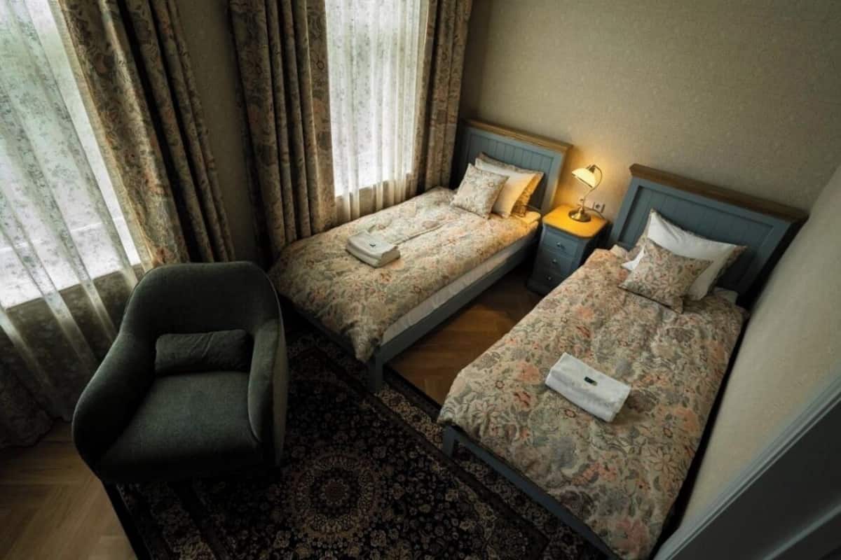 Una habitación de hotel con dos camas individuales, una silla, una mesita de noche con lámpara y una ventana con cortinas.