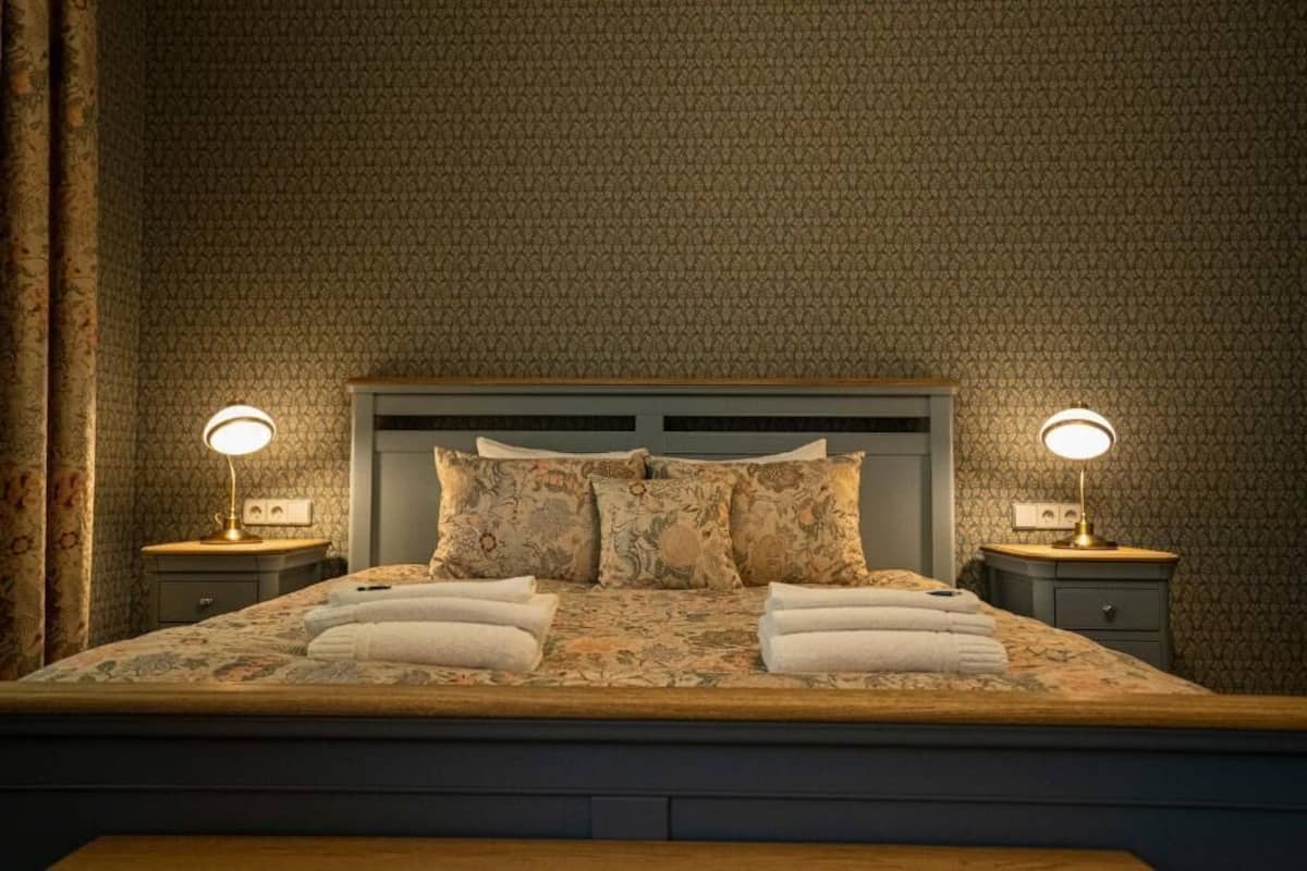 Una cama bien hecha con ropa de cama floral y dos almohadas, flanqueada por dos mesitas de noche con lámparas.