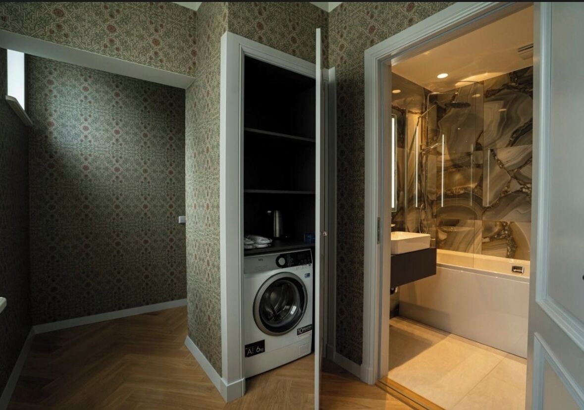A compact laundry room with a washing machine and a small shelf, adjacent to a bathroom with a marble backsplash and a bathtub.