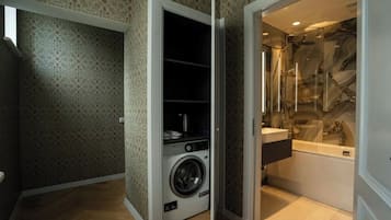 A compact laundry room with a washing machine and a small shelf, adjacent to a bathroom with a marble backsplash and a bathtub.