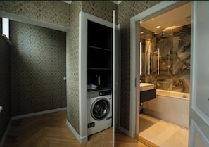 Ein kleiner Hauswirtschaftsraum mit Waschmaschine und einer kleinen Ablage, angrenzend an ein Badezimmer mit Marmorverkleidung und einer Badewanne.