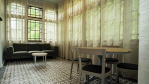 Een kamer met een patroon vloer, een bank, een ronde tafel met stoelen, en grote ramen met gordijnen.