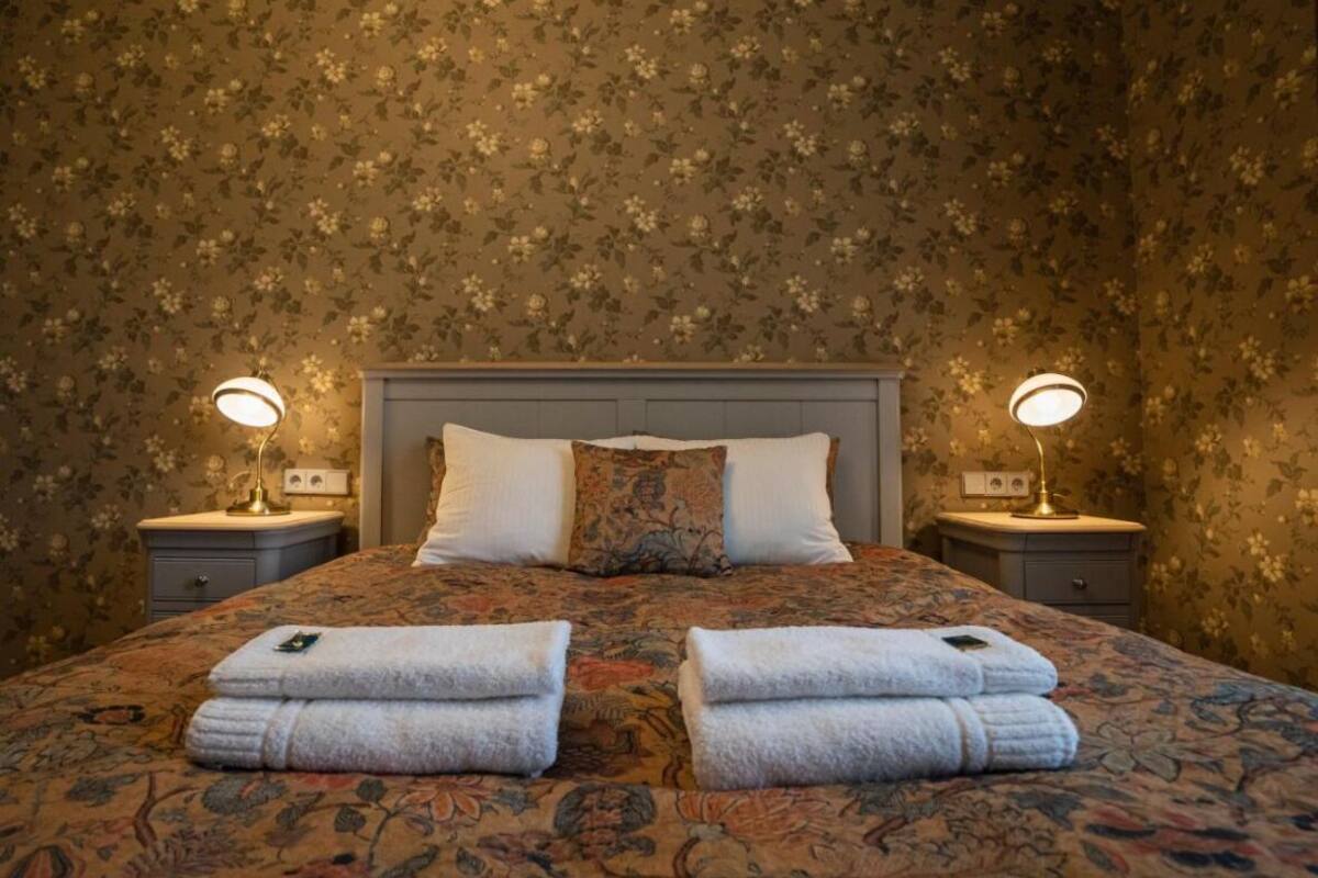 Una cama bien hecha con ropa de cama estampada con motivos florales, dos lámparas de noche y toallas dobladas sobre la cama.