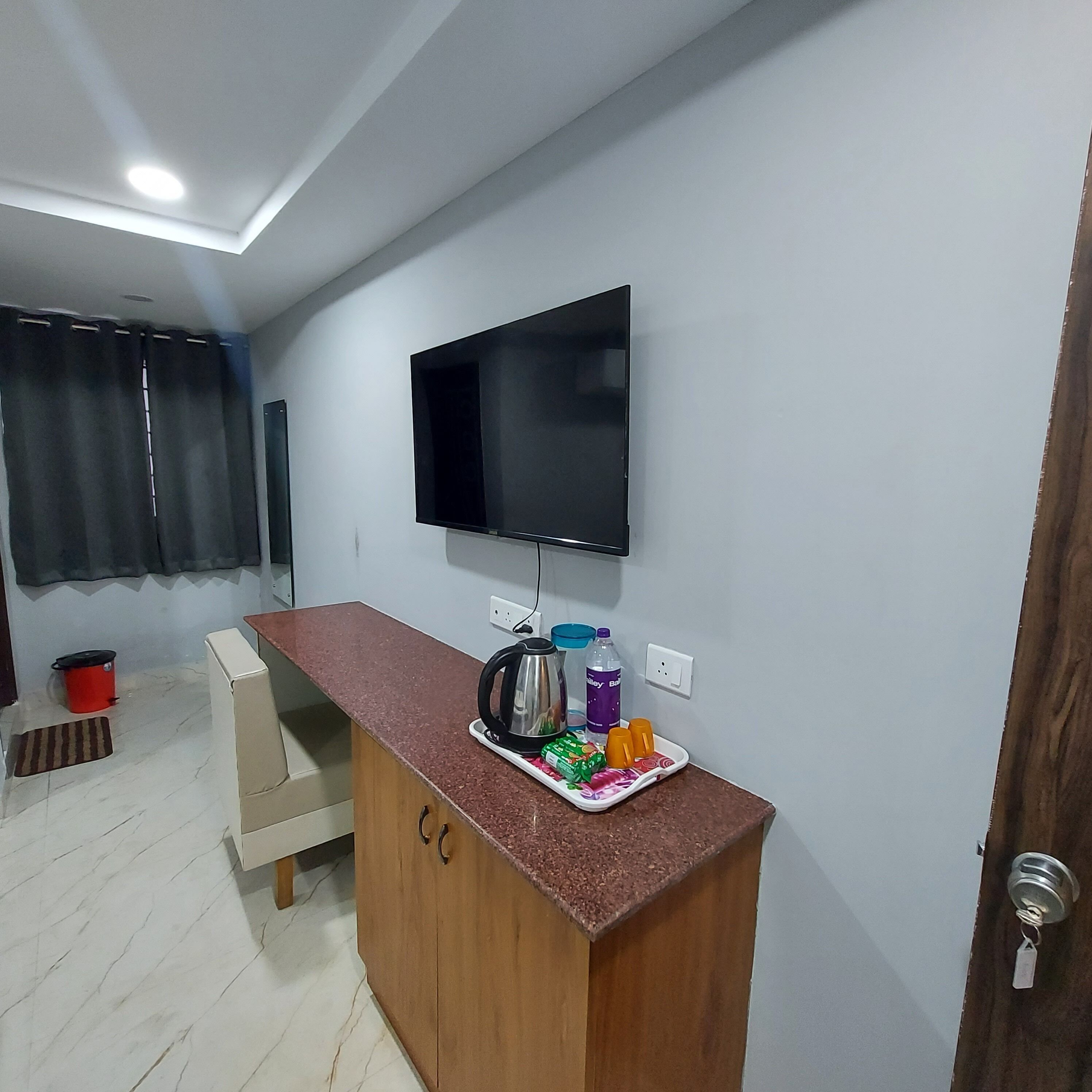 Uma pequena cozinha compacta com televisão na parede, um armário de madeira e bancada com chaleira e garrafas de água.