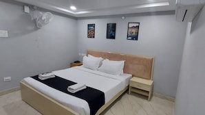 Un lit bien fait, avec du linge de lit blanc et un élément décoratif noir, une table de chevet avec une lampe, et trois tableaux encadrés accrochés au mur.