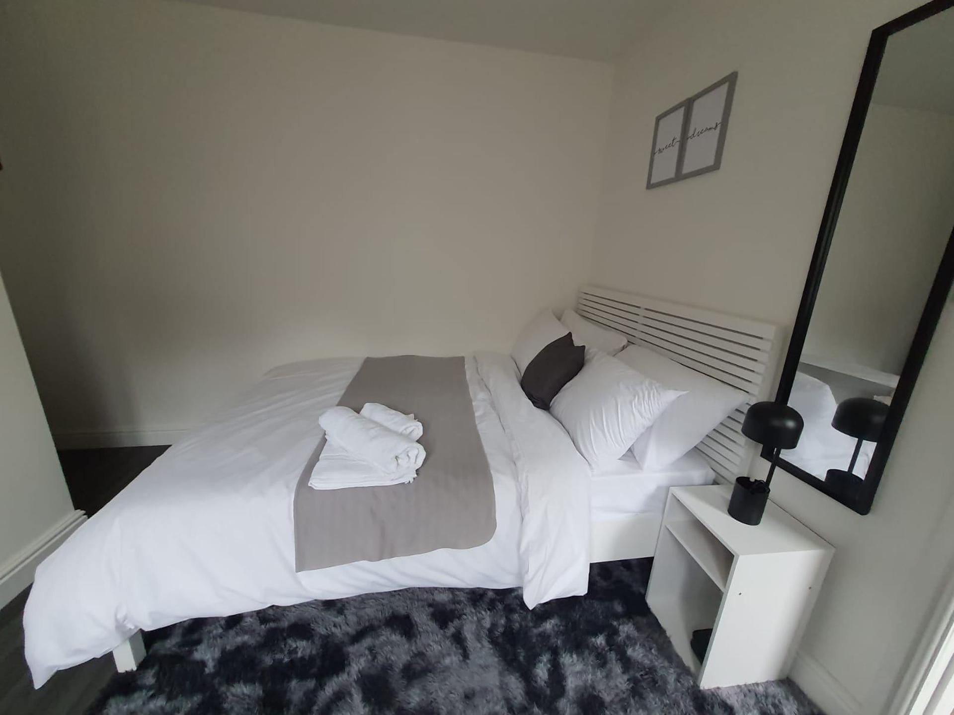A neatly made bed with white linens and a grey throw, a bedside table with a lamp, and a mirror on the wall.