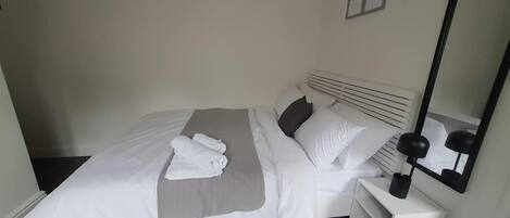 A neatly made bed with white linens and a grey throw, a bedside table with a lamp, and a mirror on the wall.