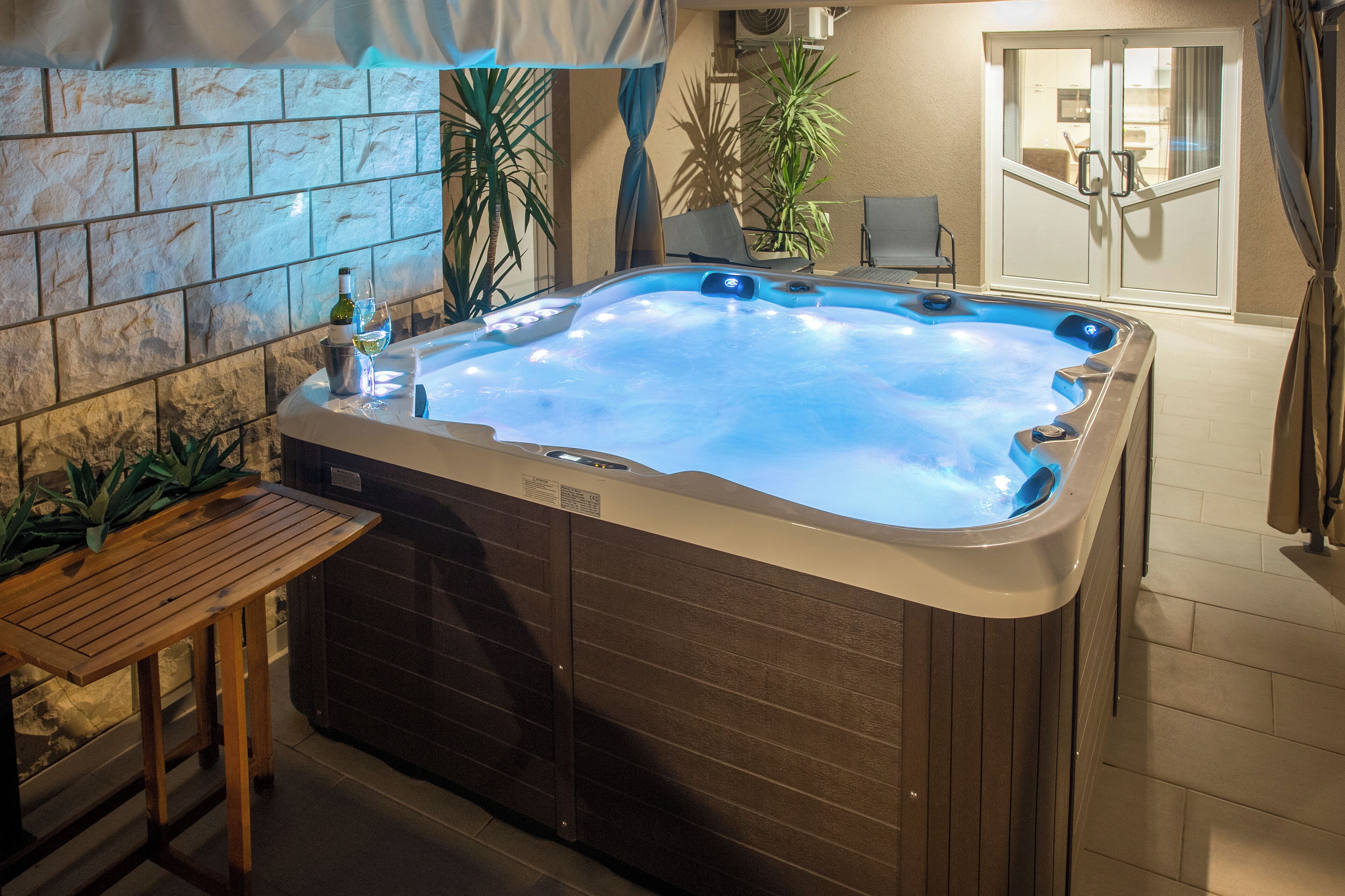 A hot tub with a wooden table and chairs nearby, surrounded by stone walls and potted plants.