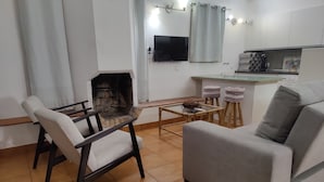 A living room with a fireplace, a sofa, a coffee table, and a kitchen area with a TV and a kettle.
