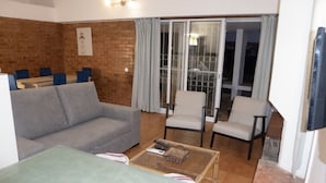A living room with a grey sofa, two armchairs, a coffee table, and a dining table with chairs. There is a sliding glass door leading to a balcony with a railing.