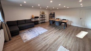 Een moderne woonkamer met een zwarte bank, een houten eettafel met stoelen, een boekenkast en inbouwspots.