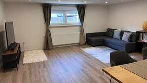 Een moderne woonkamer met een hoekbank, een flatscreen televisie, een houten salontafel en een groot raam met gordijnen.