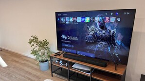 Een flatscreen televisie op een houten tv-meubel, met een afstandsbediening, een spelcontroller en een bloempot ernaast.