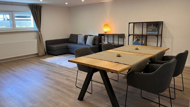Een moderne woonkamer met een houten eettafel, een grijze bank en een bijzettafeltje met een lamp.