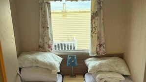 A small, cozy room with two single beds, a nightstand with a lamp, and a window with curtains. - 3 bed With Balcony on Lido Beach (Prestatyn)