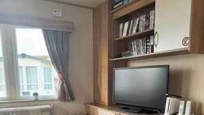 A compact RV interior with a sofa, a television, a bookshelf, and a fireplace. - 3 bed With Balcony on Lido Beach (Prestatyn)