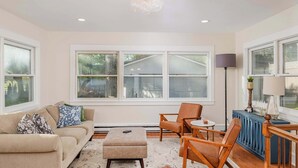A living room with a beige sofa, a coffee table, and two wooden chairs. There are large windows with a view of a house and trees. A blue radiator is visible on the right. - Historic Appleton Mansion-Sleeps 13 & Free Parking (Appleton)