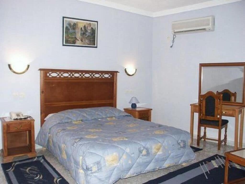 A bedroom with a wooden bed, bedside tables, a desk, a chair, a mirror, and a wall-mounted air conditioner.