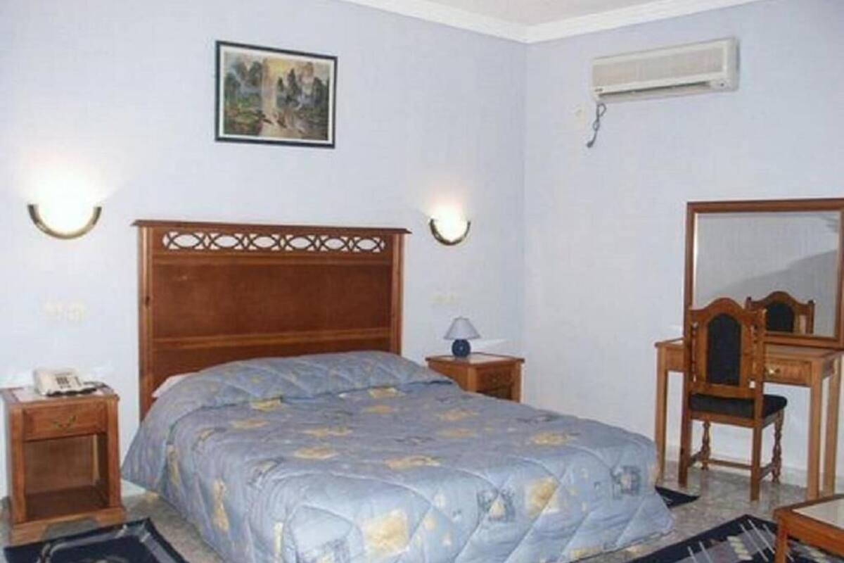 A bedroom with a wooden bed, bedside tables, a desk, a chair, a mirror, and a wall-mounted air conditioner.