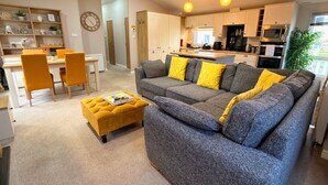 A modern living room with a grey sofa and yellow cushions, a yellow ottoman, and a dining area with a white table and yellow chairs. - Meadows 33 Hot tub - Southview Holiday Park (Skegness)