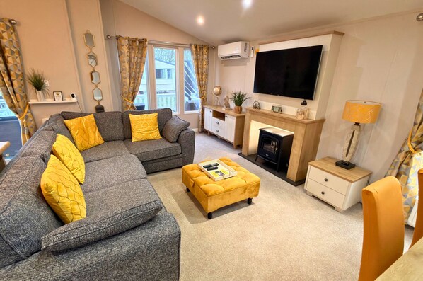 A modern living room with a sectional sofa, ottoman, TV, fireplace, and a small side table. - Meadows 33 Hot tub - Southview Holiday Park (Skegness)