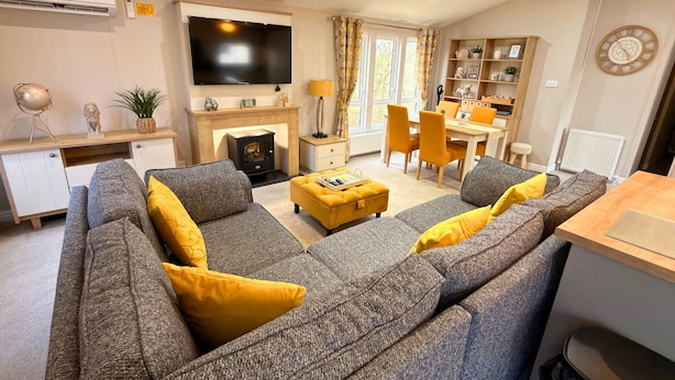 A modern living room with a grey sofa, yellow cushions, a flat-screen TV, a fireplace, and a dining area with yellow chairs. - Meadows 33 Hot tub - Southview Holiday Park (Skegness)