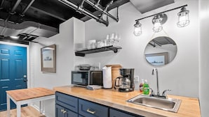 Una cucina moderna con piano di lavoro in legno, forno a microonde integrato, macchina del caffè e lavello. Al di sopra del bancone, scaffali con bicchieri e uno specchio sulla parete.