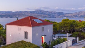 A modern white building with a red roof, a red door, and a solar panel. It is surrounded by greenery and overlooks a body of water with mountains in the background. - Luxurious apartment in Postira (Postira)