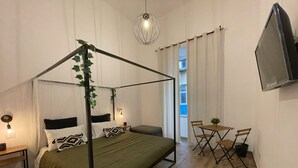 Habitación con cama con dosel, una mesita con sillas, televisor montado en la pared y una ventana con cortinas.