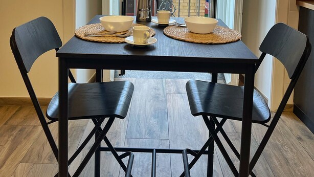 Un comedor con una mesa lista para dos personas, una ventana con vistas a edificios y una puerta a la derecha.