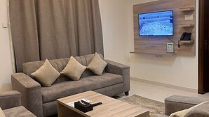 A modern living room with a grey sofa, matching armchairs, and a wooden coffee table. A flat-screen TV is mounted on the wall, and a phone is placed on a shelf. - Sweet Star Serviced Apartments (Tabuk)