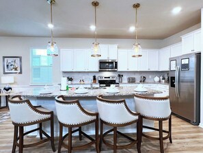 A modern kitchen with white cabinets, a stainless steel refrigerator, a microwave, and a granite countertop. There are three bar stools with wooden frames and white cushioned seats.