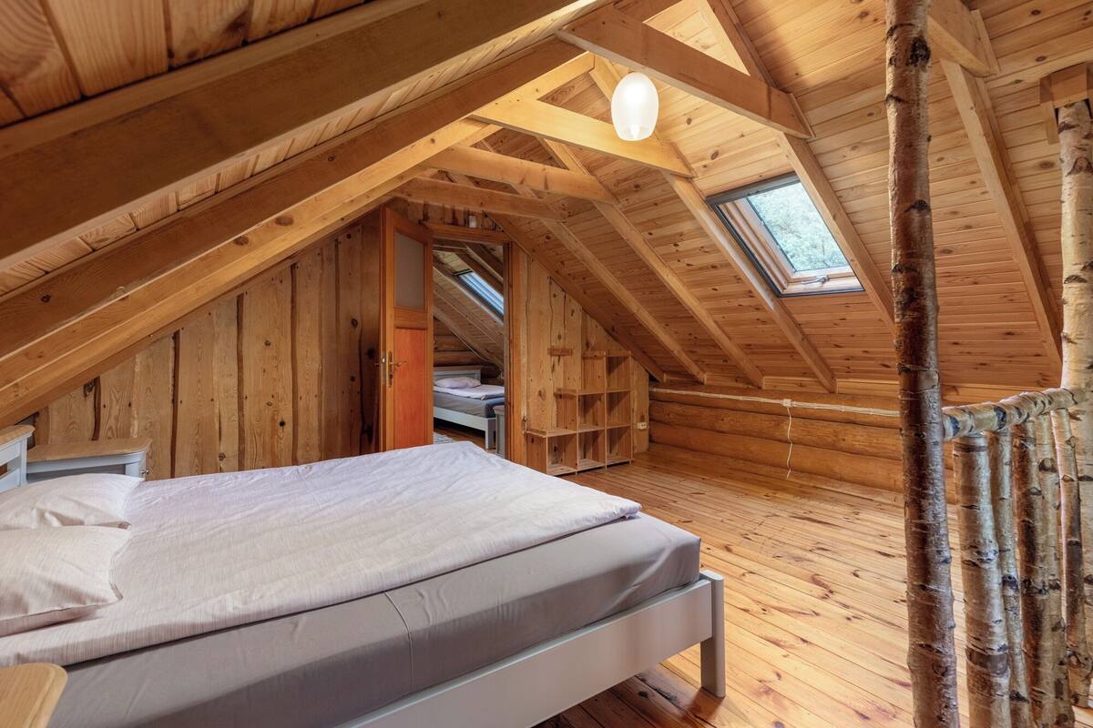 Een knus slaapkamer onder het dak, met schuine wanden, houten balken en een dakraam, en een bed met een wit matras en kussens.