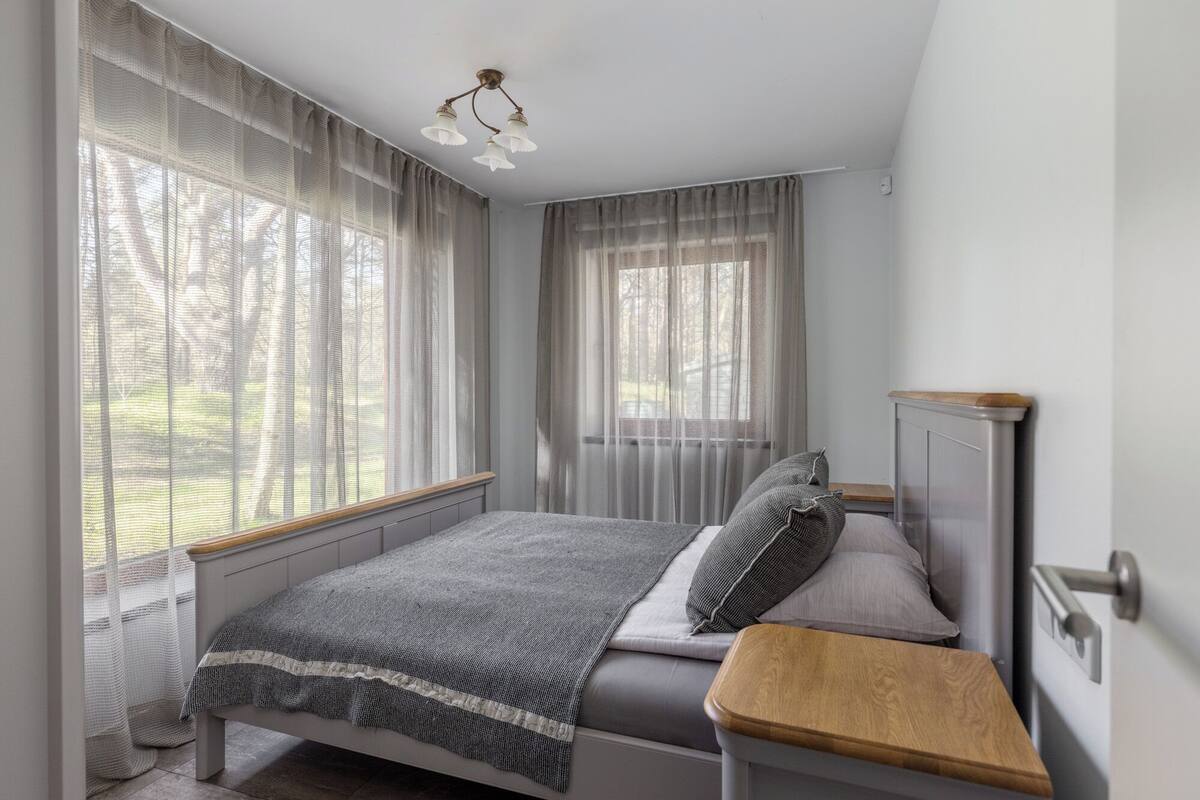 Een slaapkamer met een bed, een nachtkastje, een raam met dunne gordijnen en een plafondlamp.