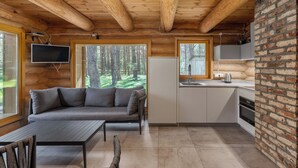 Interior de uma cabana de tronco com sofá, mesa de jantar, cozinha e uma grande janela com vista para uma floresta.