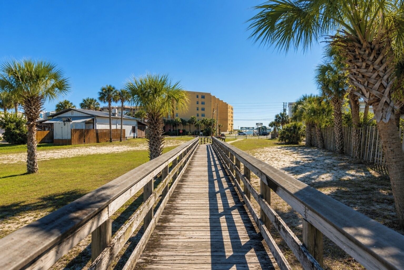 Un paseo marítimo de madera que conduce a un edificio de techo plano, rodeado de palmeras y una playa de arena.