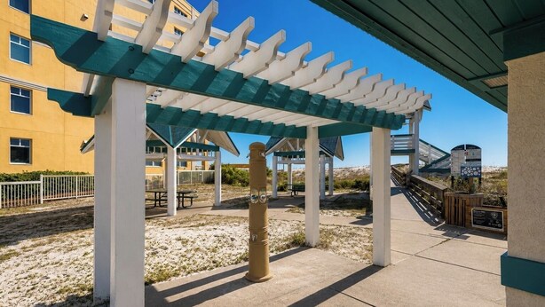A covered outdoor area with a pergola structure, a sandy ground, and a building in the background. - Tiny Home on the Boardwalk | Walk to the Beach (Fort Walton Beach)