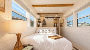A bedroom with a large bed, a bedside lamp, a bedside table with a book and a decorative item, and a window with a view of the outside. - Tiny Home on the Boardwalk | Walk to the Beach (Fort Walton Beach)