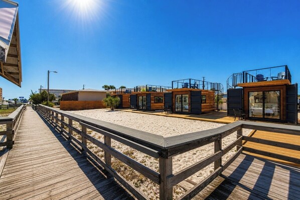 Une promenade en bois mène à une propriété située en bord de mer, comprenant plusieurs cabanes et une pièce entièrement vitrée.