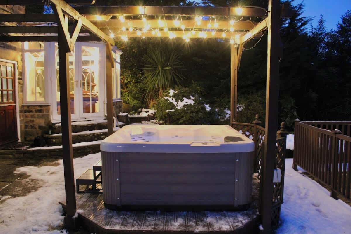 Een jacuzzi onder een houten pergola met lichtslingers, omringd door sneeuw en een stenen gebouw met ramen.
