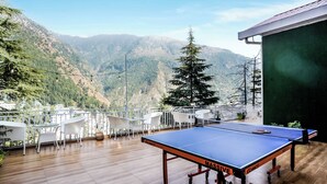 Mesa de ping pong sobre una terraza de madera con sillas blancas, con vistas a un paisaje montañoso.