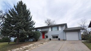 A two-story house with a grey exterior, a red front door, and a garage. There are two large potted plants in front of the house, and a large evergreen tree to the left.